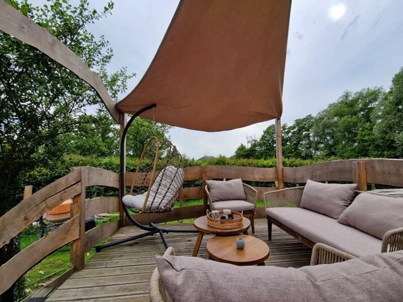 Terrasse en bois avec canapé, fauteuil suspendu et table basse sous un grand voile d'ombrage beige.