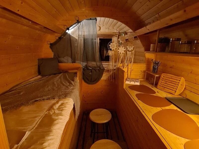Intérieur en bois d'une petite chambre avec lit, tabourets, rideau noir et décorations bohèmes suspendues.