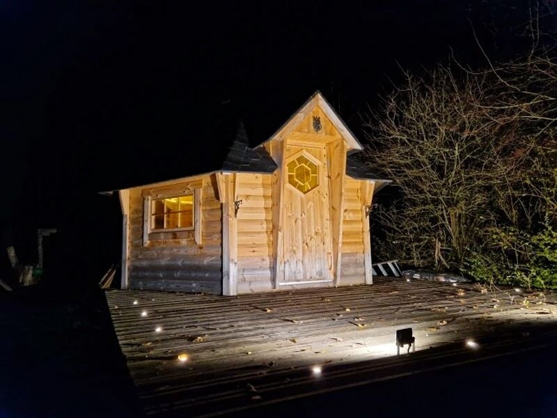 Petite cabane en bois éclairée la nuit, entourée d'arbres, avec une porte centrale et un toit pointu.
