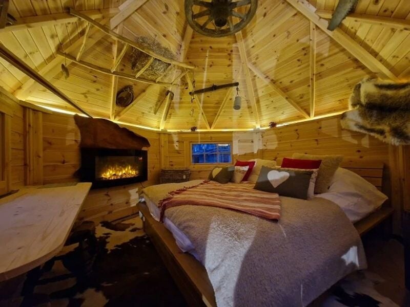 Intérieur chaleureux d'une cabane en bois avec un lit, cheminée allumée, décor rustique et poutres apparentes.