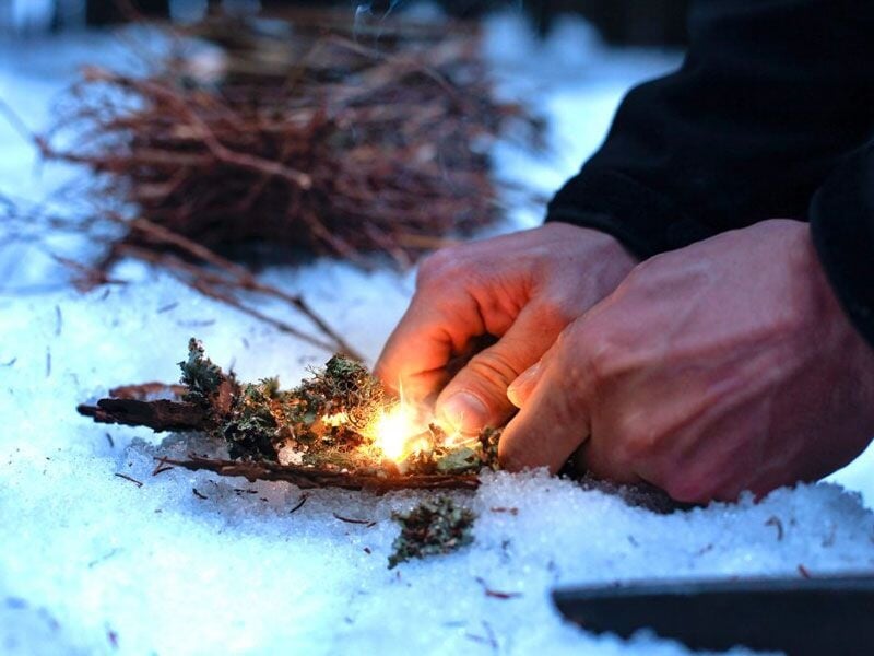 Une personne allume un feu avec des brindilles et de la mousse sur un sol enneigé en stage de survie.