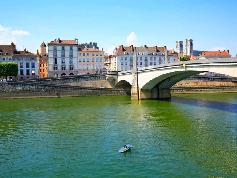 Vue d'un pont sur une rivière avec un petit bateau et des bâtiments anciens en arrière-plan sous un ciel bleu.