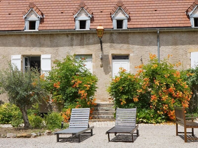 Façade d'une maison en pierre avec deux chaises longues devant un jardin fleuri et un lampadaire ancien.