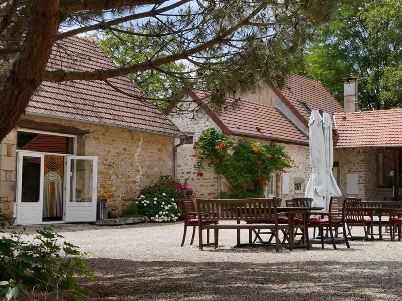 Cour extérieure avec tables, chaises en bois et parasol fermé devant une maison en pierre sous des arbres.