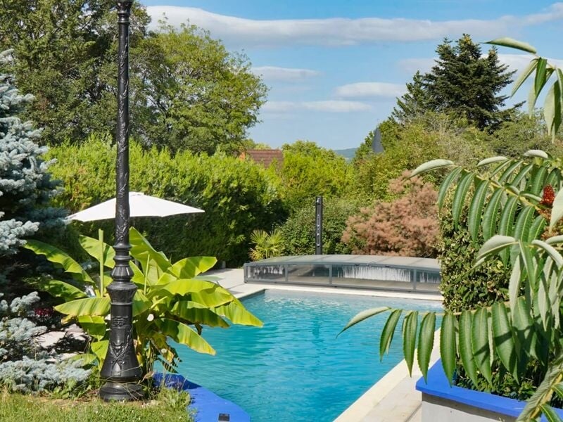 Piscine bleue entourée de végétation luxuriante et d'un lampadaire noir dans un jardin ensoleillé.