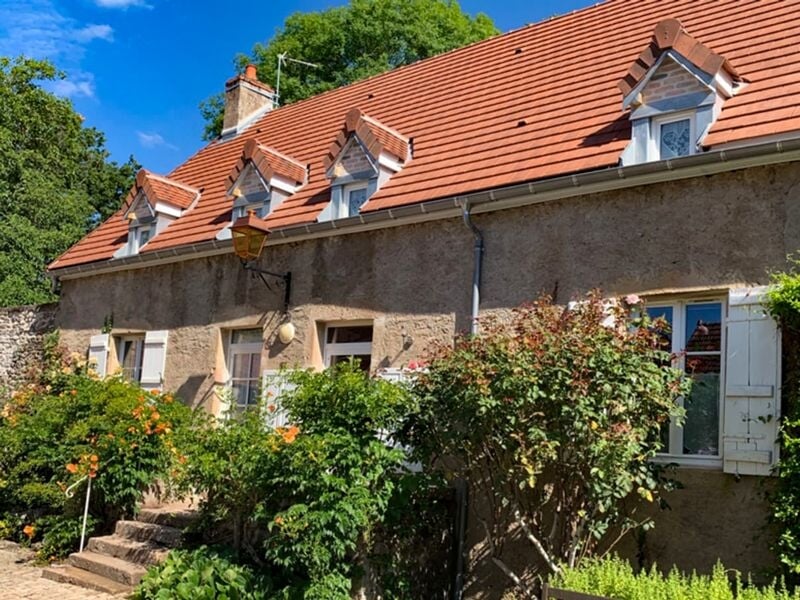 Façade d'une maison ancienne avec toit en tuiles rouges, fenêtres blanches et buissons fleuris au premier plan.