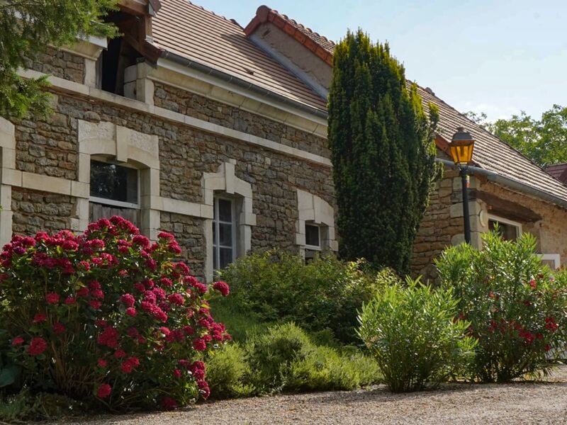 Maison en pierre avec fenêtres à volets blancs, entourée de buissons verts et de fleurs rouges sous un ciel bleu.