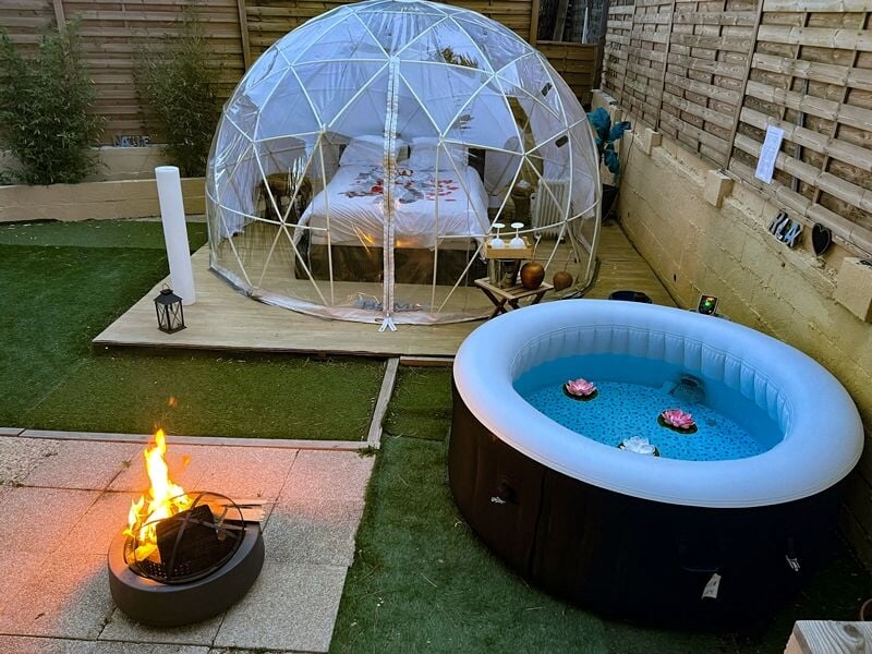 Dôme transparent dans un jardin avec un lit à l'intérieur, un jacuzzi gonflable et un feu de camp allumé à côté.