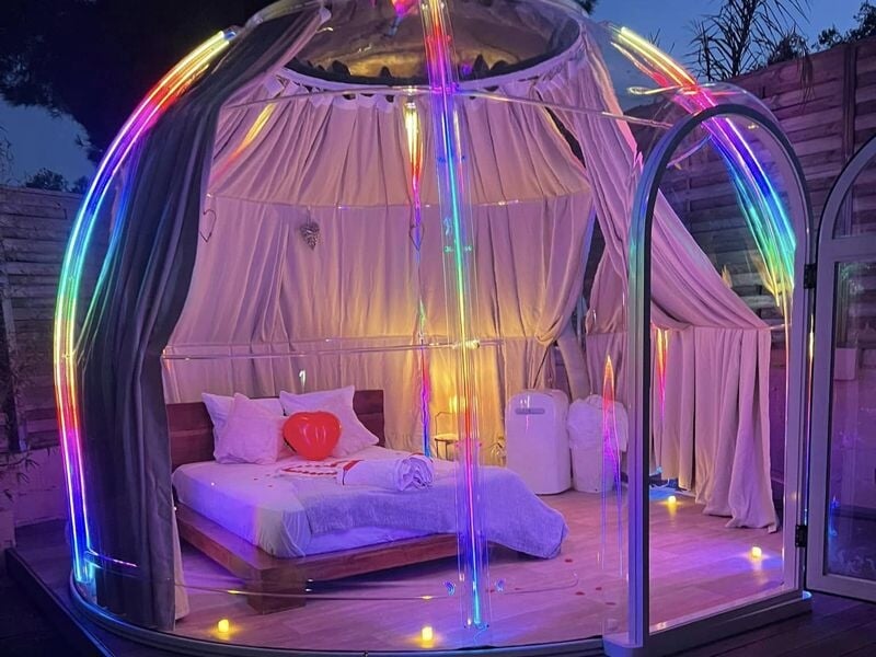 Intérieur d'un dôme lumineux avec un lit double, des rideaux blancs et des lumières colorées en arc-en-ciel.