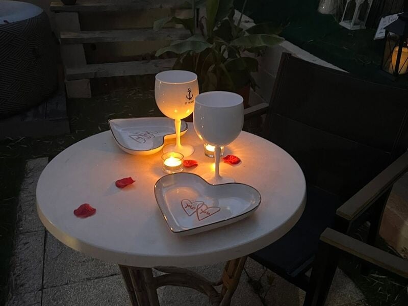 Table ronde avec deux verres blancs, bougies allumées et pétales de rose pour une ambiance romantique en soirée.