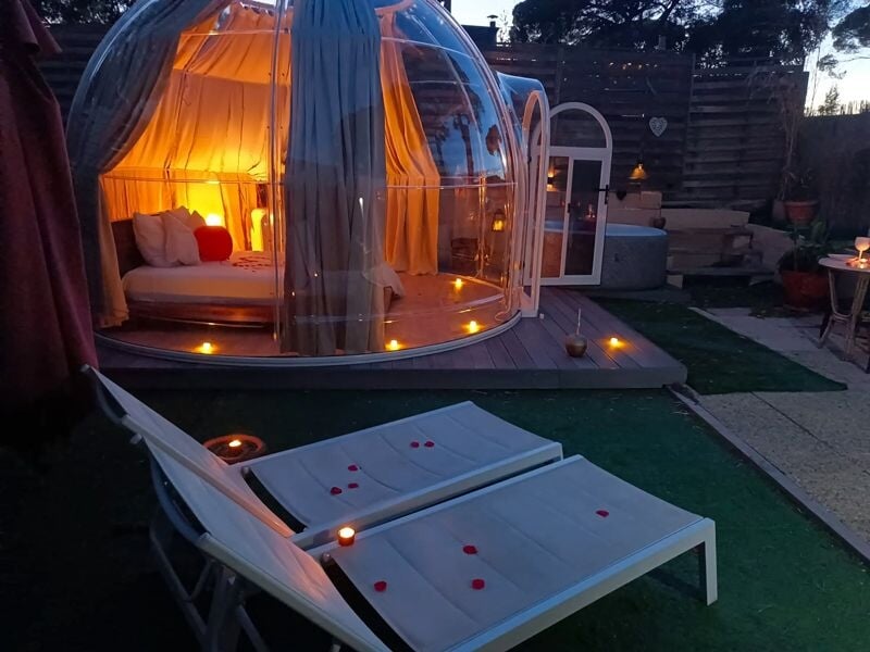Dôme éclairé avec un lit à l'intérieur, chaises longues blanches et ambiance romantique avec des pétales de rose.