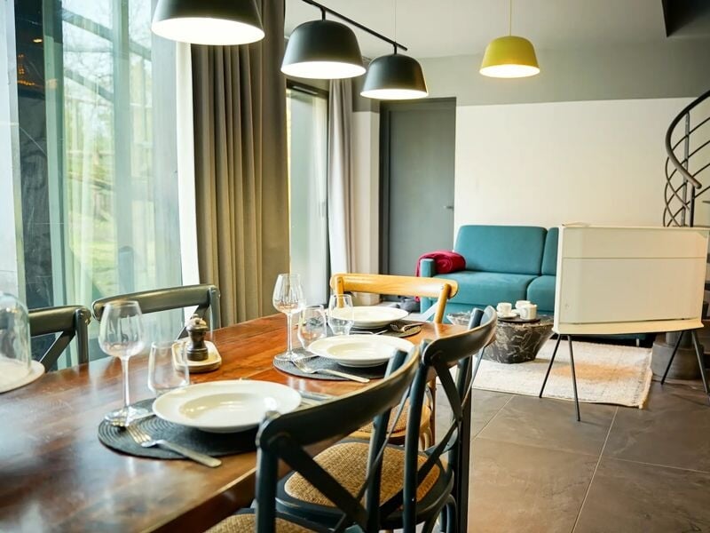 Salle à manger moderne avec table en bois, chaises variées et grandes fenêtres laissant entrer la lumière naturelle.