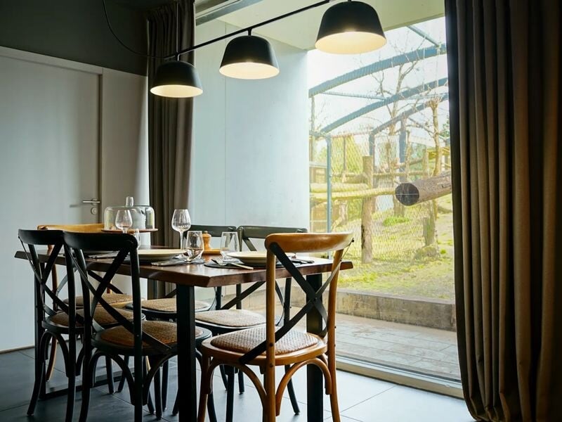 Table à manger en bois avec six chaises, éclairage suspendu et vue sur un jardin extérieur à travers une baie vitrée.