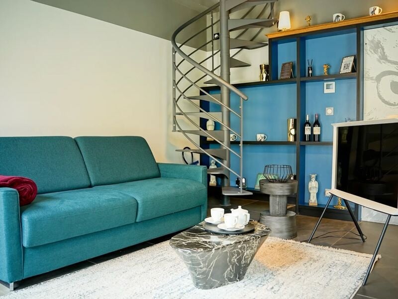 Salon cosy avec canapé bleu, table basse en marbre, escalier en colimaçon et étagères décorées.