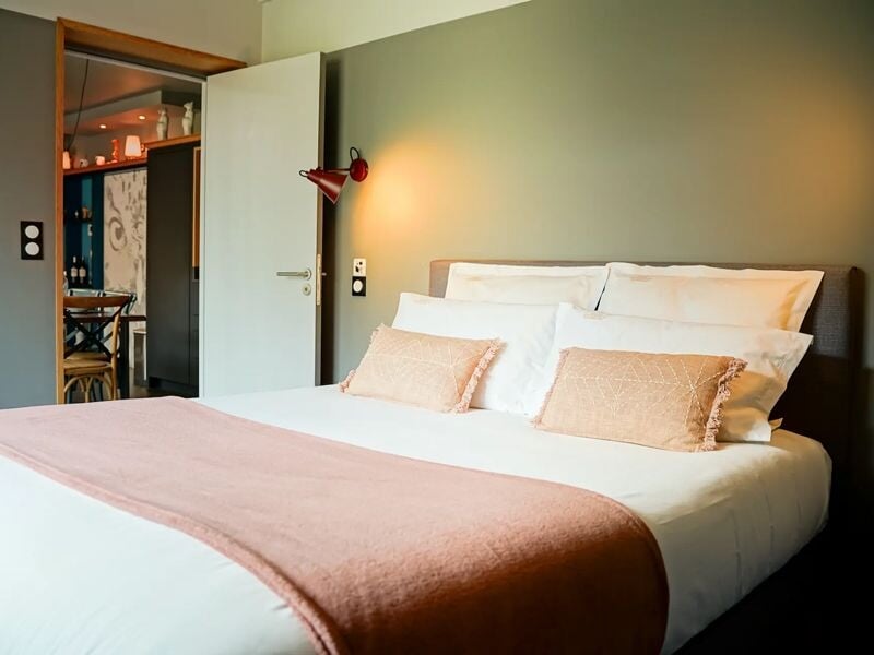 Chambre élégante avec lit double, linge blanc, couverture rose et lampe murale rouge près de la porte ouverte.