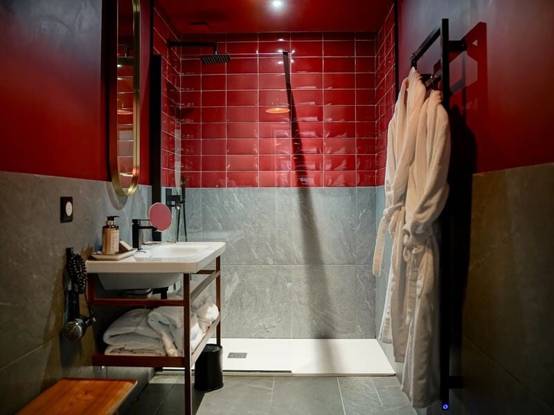 Salle de bain moderne avec douche à l'italienne, carrelage rouge et gris, lavabo et peignoirs suspendus.