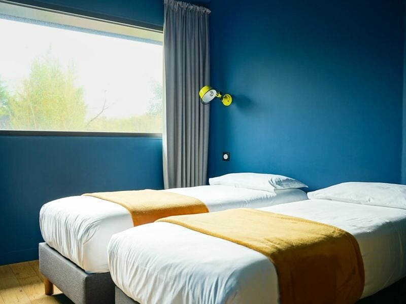 Chambre avec deux lits simples, murs bleus, rideaux gris et lampes murales jaunes au-dessus des lits.