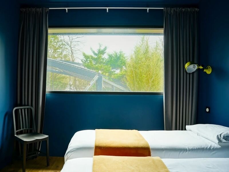 Chambre double avec deux lits simples, grande fenêtre, murs bleus et rideaux gris foncés.