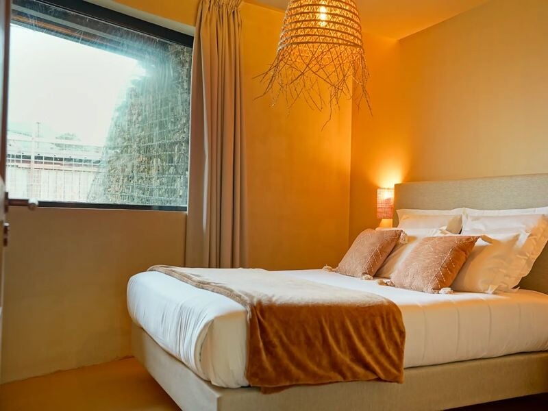Chambre chaleureuse avec lit double, coussins beige et couverture marron, lumière douce et grande fenêtre.
