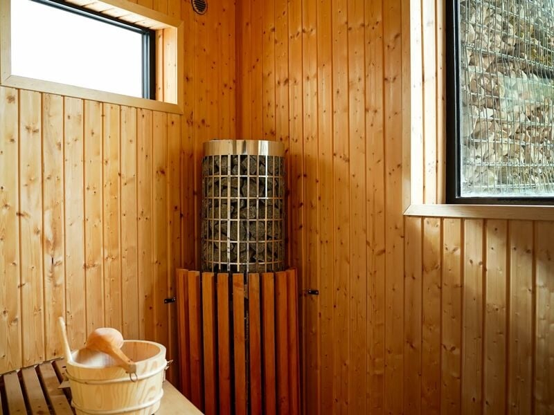 Sauna en bois avec poêle à pierres, seau en bois et grande fenêtre offrant une vue extérieure.