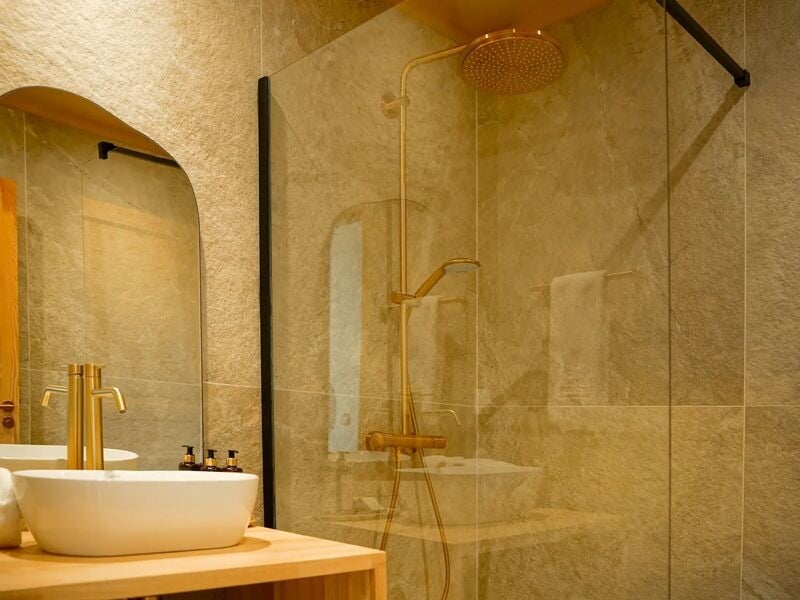 Salle de bain moderne avec douche à l'italienne, robinetterie dorée et lavabo blanc sur meuble bois.