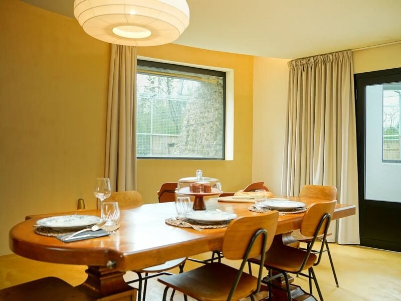 Salle à manger avec table en bois dressée, chaises vintage et grande fenêtre avec rideaux beiges.