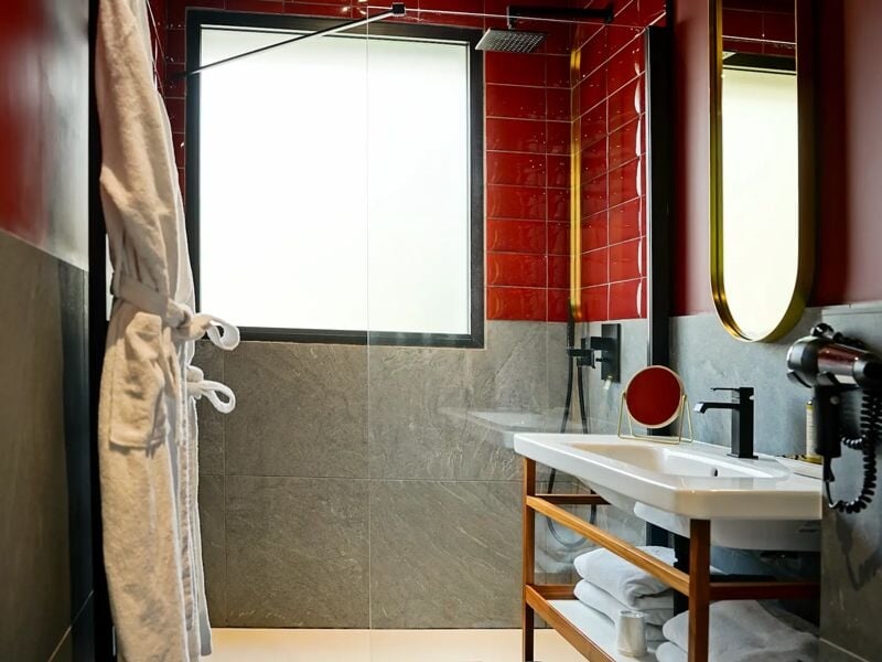 Salle de bain avec douche à l'italienne, murs rouges et gris, fenêtre et peignoir suspendu sur le côté.