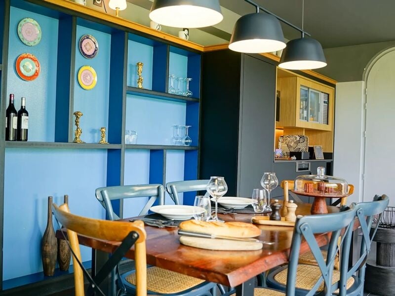 Salle à manger avec table en bois, chaises colorées, vaisselle et décorations sur étagères bleues au mur.