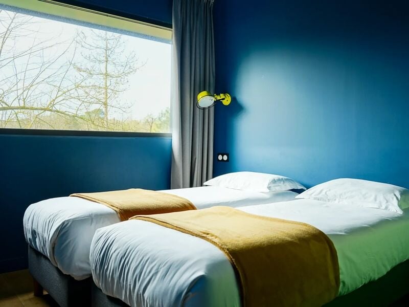 Chambre avec deux lits simples, murs bleus, grandes fenêtres et couvertures jaunes sur les lits.