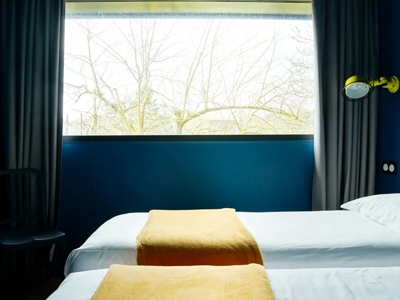 Vue latérale d'une chambre avec deux lits simples, murs bleus et grandes fenêtres laissant entrer la lumière.