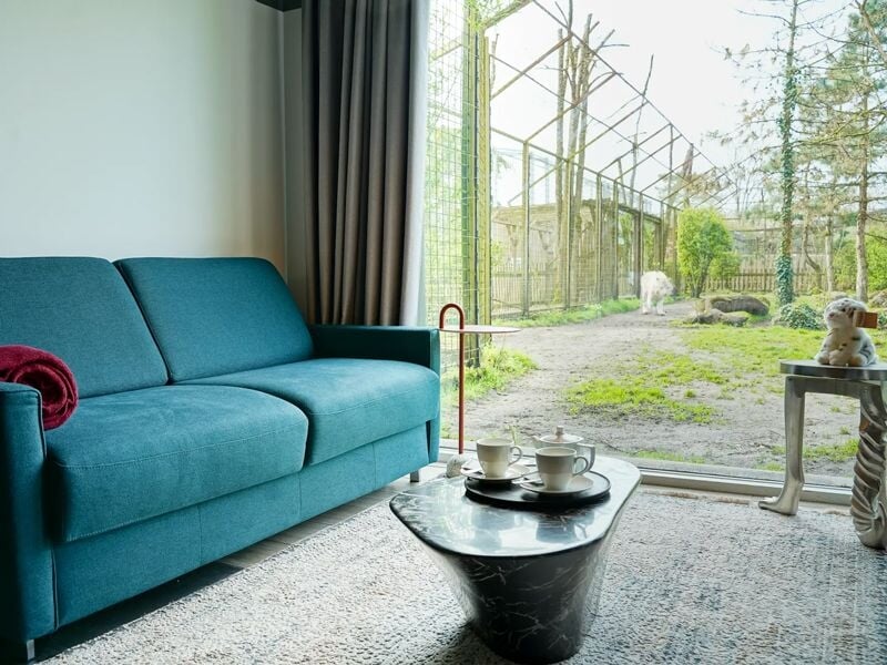 Salon avec canapé bleu, table basse noire et grande baie vitrée donnant sur un jardin avec animaux.