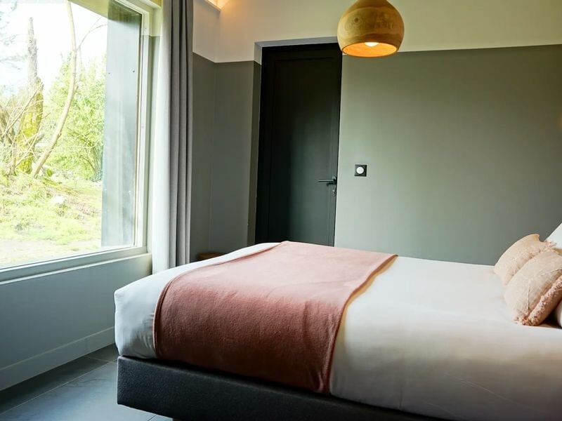 Autre angle d'une chambre avec lit double, murs verts et couverture rose, éclairée par une fenêtre.