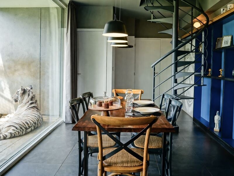 Salle à manger avec table en bois, chaises noires, escalier en colimaçon et grande baie vitrée avec tigre blanc.