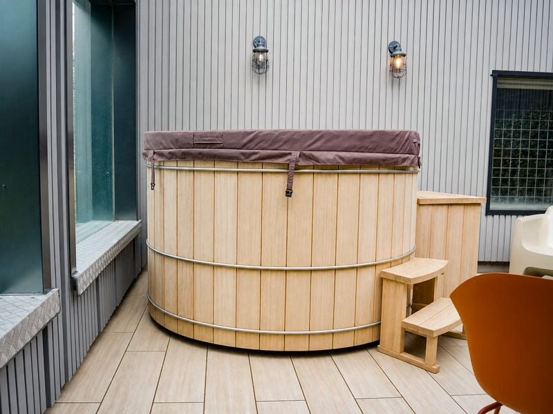 Vue rapprochée d'un spa en bois sur une terrasse avec chaises colorées et mur gris clair en arrière-plan.