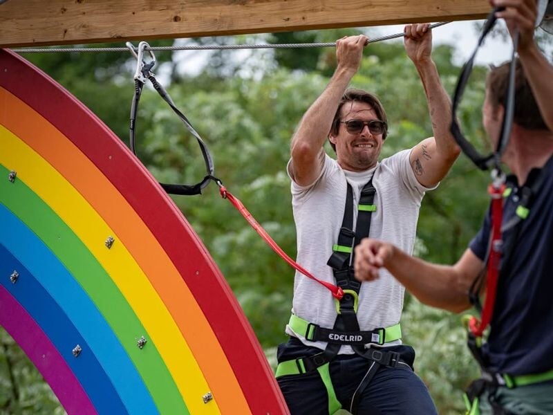 Homme souriant équipé d'un harnais de sécurité accroché à une structure colorée en forme d'arc-en-ciel en accrobranche.