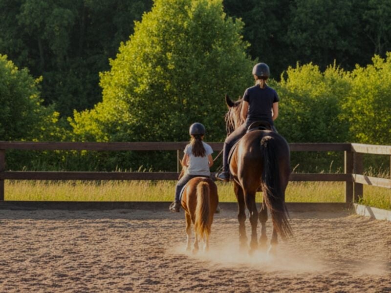 Deux cavaliers, un adulte et un enfant, montent leurs chevaux dans une carrière en plein air au coucher du soleil.