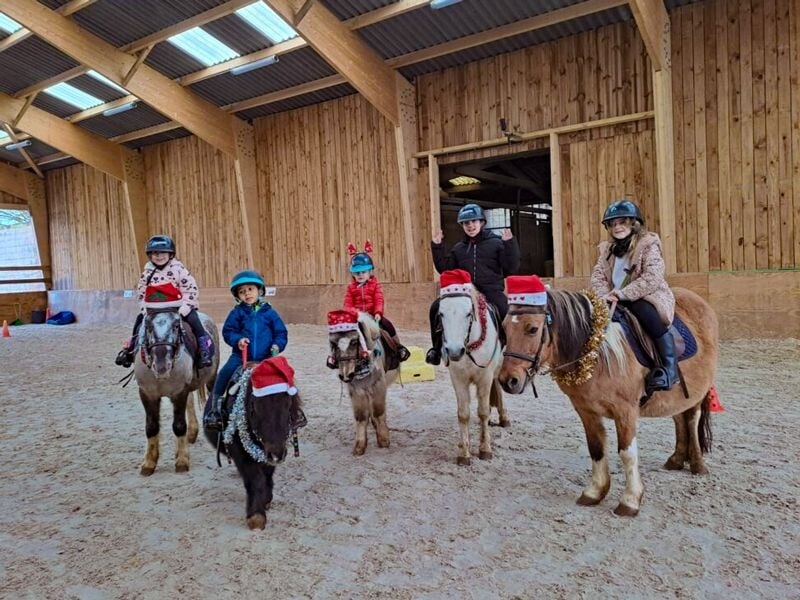 Groupe d'enfants sur des poneys décorés de bonnets de Noël dans une écurie en bois bien éclairée.
