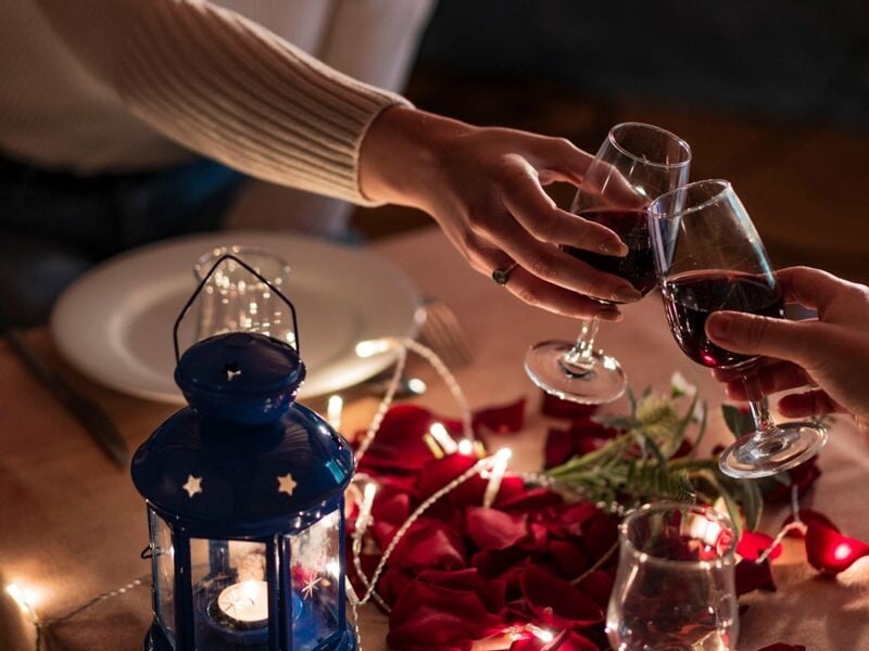 Gros plan sur deux mains qui trinquent avec des verres de vin rouge sur une table décorée de pétales et d'une lanterne bleue.