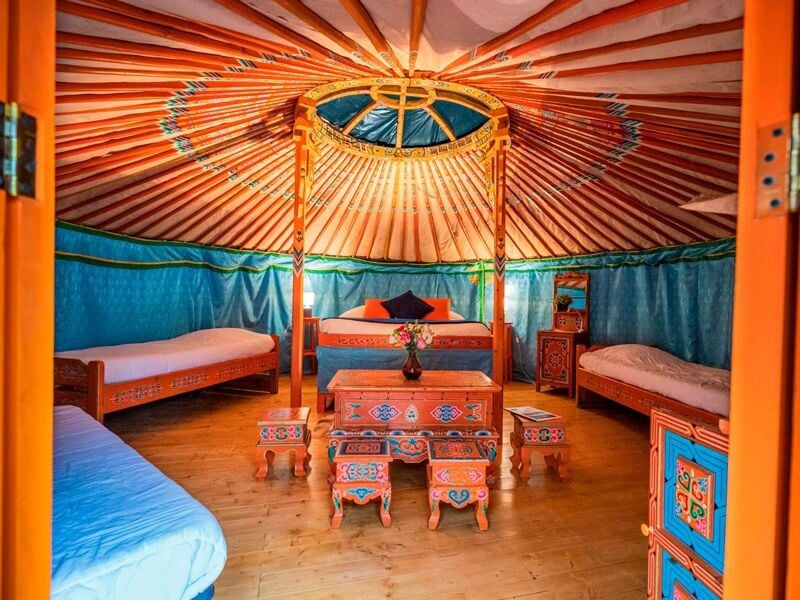 Vue intérieure d'une yourte avec un lit double, deux lits simples, un meuble peint et un plafond rond coloré.