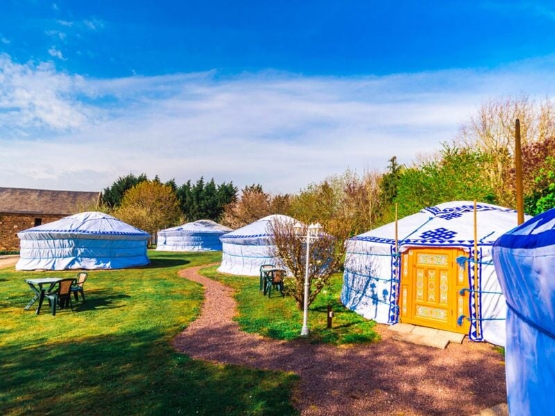 Plusieurs yourtes blanches avec des motifs bleus installées dans un jardin sous un ciel bleu avec des arbres autour.