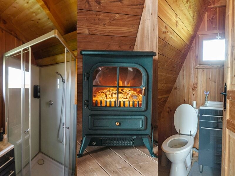 Salle de bain en bois avec douche, poêle à bois allumé et toilettes dans une petite cabane chaleureuse.