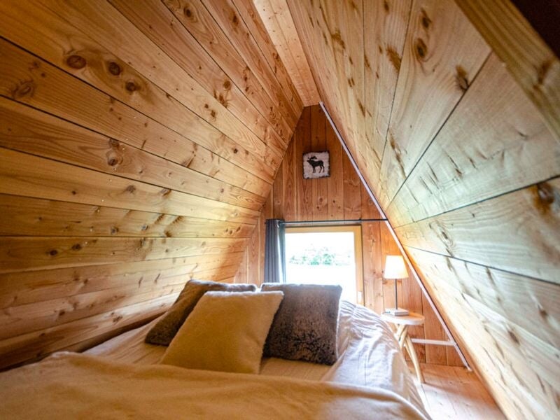 Chambre mansardée en bois avec lit double, coussins et lampe sur table de chevet près d'une fenêtre.