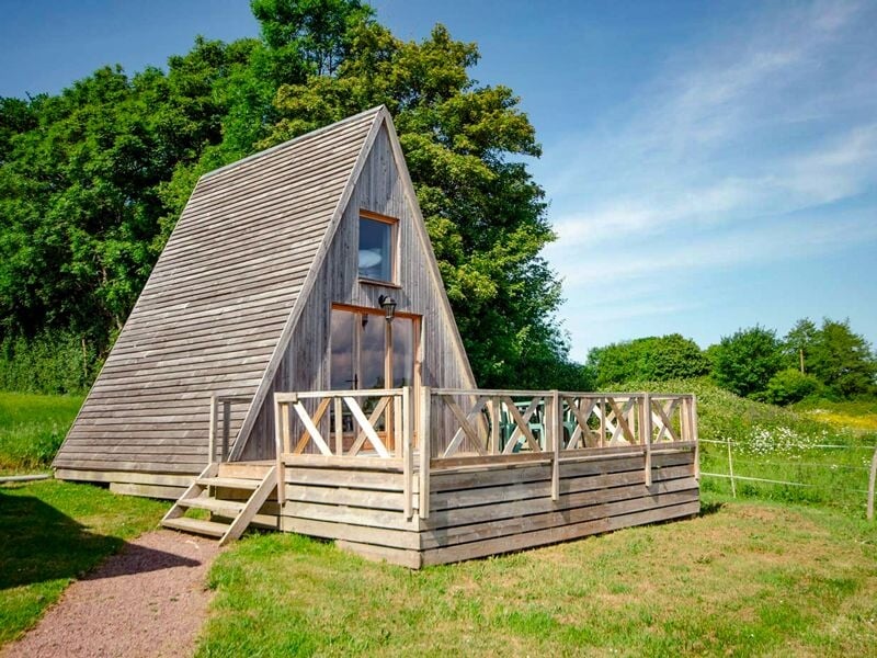 Chalet tipi en bois de forme triangulaire avec terrasse en bois, entourée d'herbe et d'arbres verts.