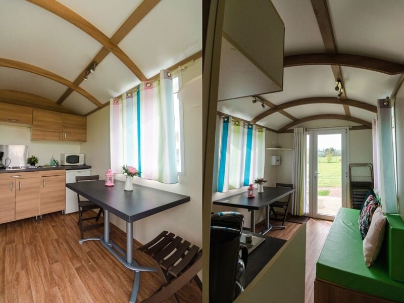 Intérieur d'une roulotte avec cuisine équipée, table noire, chaises et canapé vert, fenêtres avec rideaux colorés.