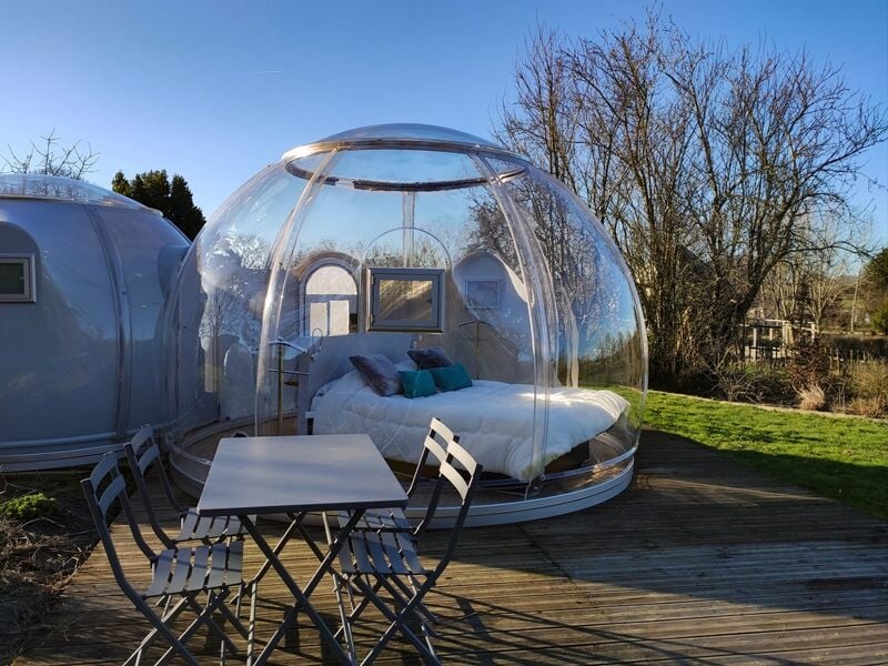 Bulle transparente avec un lit à l'intérieur et une table avec quatre chaises sur une terrasse en bois.