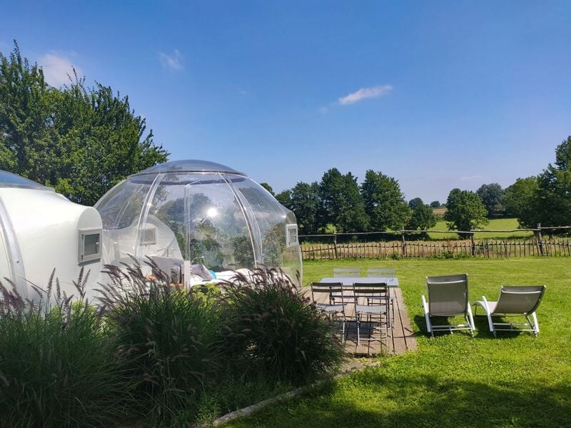 Bulle transparente avec un lit à l'intérieur et une terrasse avec table, chaises et deux transats dans un jardin.