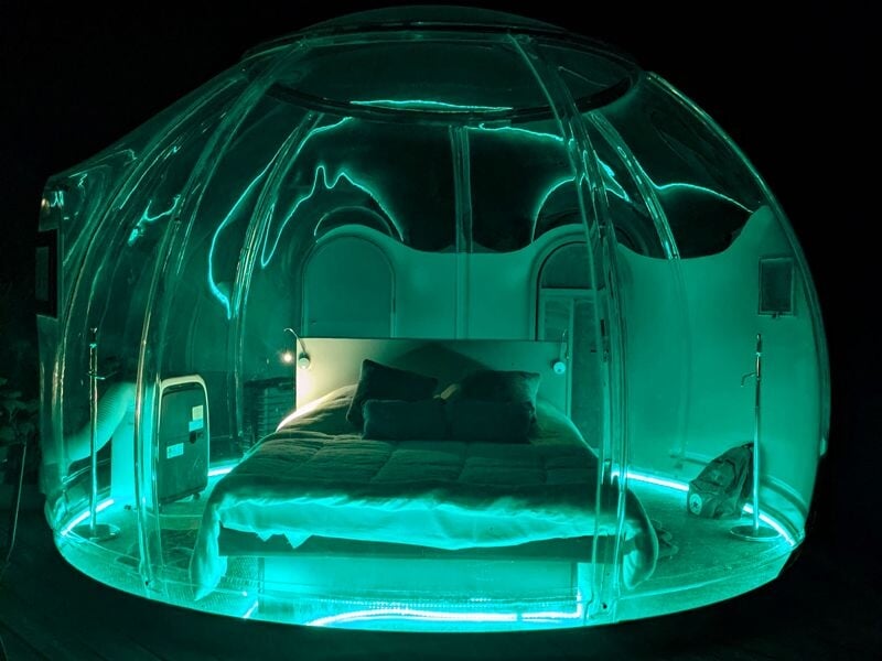 Bulle transparente éclairée en vert la nuit avec un lit et des coussins à l'intérieur.