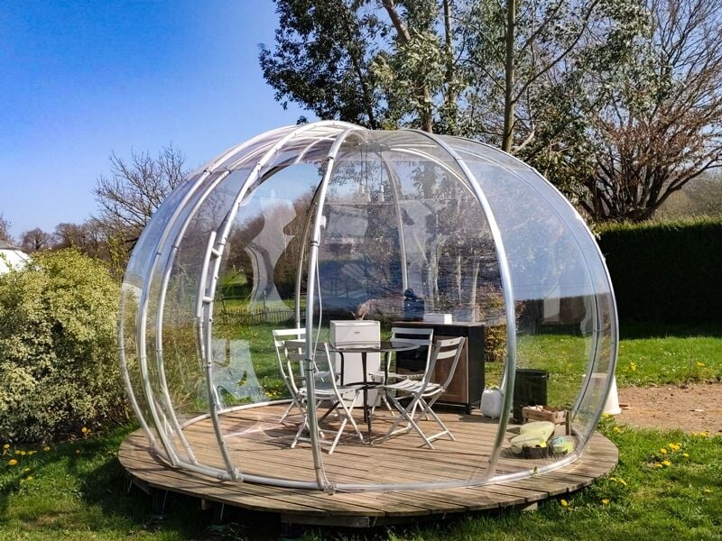Bulle transparente ronde avec une table et quatre chaises sur une terrasse en bois dans un jardin.