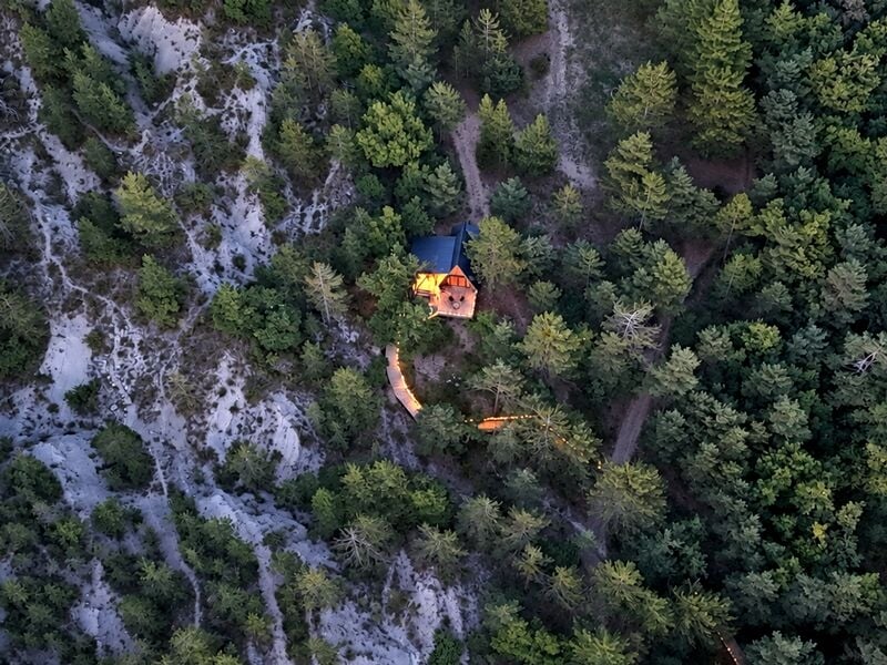 Vue aérienne d'une petite cabane en bois entourée d'arbres verts et d'un terrain rocheux en pente.