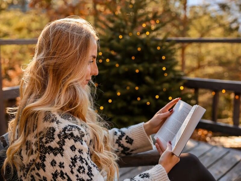 Femme blonde en pull à motifs noirs lisant un livre sur une terrasse en bois avec un sapin décoré.